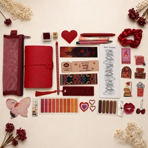 May include: A selection of red stationery items, including a journal, pens, bookmarks, and stickers, arranged on a cream-coloured surface. The arrangement includes a scrunchie and various decorative elements.