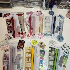 Peut inclure: Une collection de trackers de livres, d'autocollants et de stylos. Les trackers présentent des motifs d'étagères et du texte comme "Book Lover" et "Read More Books". Les stylos sont de différentes couleurs, dont le rose, le jaune et le vert. Les autocollants ont des phrases comme "Good Vibes Only."