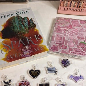 May include: A selection of book-related charms with quotes such as "Give Me More Books" and "Fantasy Please." The image also shows a book titled "Spark of Evermore" and a pink book organiser.