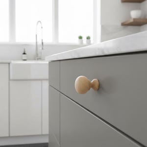 May include: A light-coloured wooden cabinet knob with a rounded design is attached to a grey cabinet door. The knob has a circular shape and a smaller, cylindrical base. The kitchen setting includes a white countertop and a window.