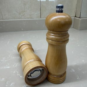 May include: Two wooden pepper grinders. One is upright, the other is lying on its side. Both are made of light-colored wood with a bulbous top and a metal grinding mechanism at the base. The grinders are designed for kitchen use.