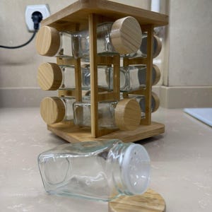 Könnte beinhalten: Ein hölzernes Gewürzregal mit zwölf Gläsern, jedes mit einem Holzdeckel. Ein Glas liegt auf der Seite, der Deckel ist abgenommen. Das Regal ist aus hellem Holz gefertigt und zur Aufbewahrung von Gewürzen bestimmt.