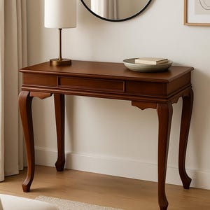 May include: A mahogany console table with a drawer and elegant curved legs. A small lamp with a cream shade sits on the table, along with a decorative bowl and books. A round mirror hangs on the wall above the table.