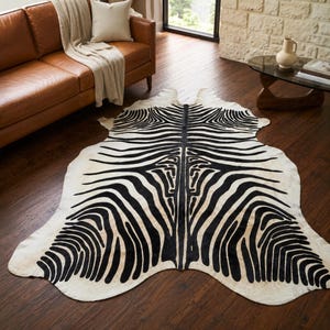 May include: A large, irregularly shaped area rug featuring a high-contrast black and white zebra stripe pattern. The rug has a natural hide-like silhouette with organic, curved edges and a dense, repeating pattern of dark stripes on a light background.