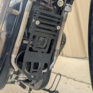 May include: Black metal motorcycle radiator guard with a slotted design, mounted on a motorcycle engine. The guard is secured with bolts and protects the radiator from debris. The engine components include silver hoses and fittings.
