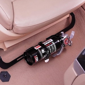 May include: A black fire extinguisher with silver accents, mounted in a vehicle. The extinguisher is secured by a black bracket and is positioned near a beige car seat. The extinguisher has the text "HR GUARD" and "13R".