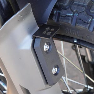 May include: Close-up of a motorcycle wheel with a black tire and silver spokes. A black metal bracket is attached to the gray fender with silver bolts. The tire has the word "RIDE" printed on it.