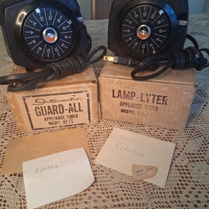Peut inclure: Deux minuteries d'appareils noires avec des cadrans circulaires, des cordons et des boîtes. Les cadrans affichent les heures de 2h à 12h. Les boîtes sont étiquetées "Intermatic GUARD-ALL" et "LAMP-LYTER". Des notes manuscrites indiquent "Kitchen".