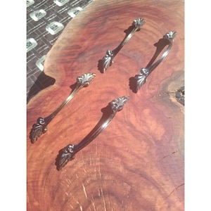Puede incluir: Cuatro tiradores de cajón de metal curvado con acabado metálico y detalles ornamentales en forma de hoja en cada extremo. Los tiradores están dispuestos en formación escalonada, mostrando una superficie pulida que refleja la luz.