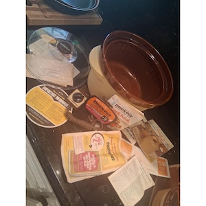 Puede incluir: Una olla de cocción lenta Crock-Pot vintage con exterior color crema y recipiente interior de cerámica marrón oscuro. El conjunto incluye una tapa de cristal transparente, cable de alimentación extraíble, mando de control y varios manuales originales.