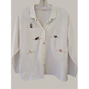May include: A white long-sleeve button-down cardigan featuring a collar and small embroidered motifs of dogs and various objects scattered across the front. The garment has a central vertical row of silver-toned metal snap buttons.
