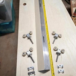 Può includere: Un insieme di componenti hardware per un sistema di porte scorrevoli. Include una guida beige e argento, quattro gruppi di rulli e due staffe metalliche. Un metro a nastro giallo è posizionato accanto alla guida.