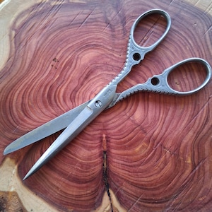 May include: Silver-colored scissors with ornate handles, resting on a reddish-brown wooden surface. The scissors have a pointed blade and a decorative design on the handles. The wood grain is visible.