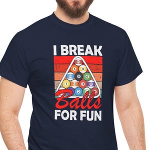 Puede incluir: Camiseta azul marino con un gráfico que presenta un triángulo de bolas de billar y el texto "I Break Balls For Fun" en rojo y blanco. El diseño está sobre un fondo retro a rayas naranjas y rojas.