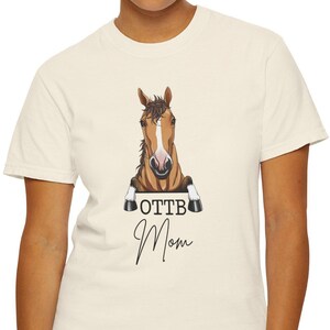 May include: Cream-colored t-shirt featuring a horse graphic with the text "OTTB Mom" in black script. The horse graphic is a detailed illustration of a brown and white horse head. The shirt is a casual, everyday wear item.