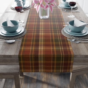 May include: A rectangular table runner featuring a plaid pattern in shades of rust orange, deep red, olive green, and mustard yellow. The fabric runner is centred on a wooden table, topped with a clear glass vase containing pink lily flowers.
