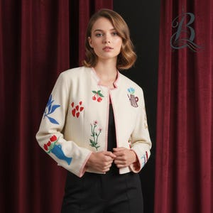 Peut inclure: Veste crème brodée avec un col rond et un intérieur noir. La veste présente des motifs floraux et en forme de cœur colorés, avec une bordure rouge et rose. Les manches sont ornées de fleurs brodées et d'un vase bleu.
