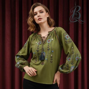 May include: Olive green long-sleeved blouse with floral embroidery in blue and brown. The blouse has a drawstring neckline and a decorative tassel. The sleeves are gathered at the wrists. The blouse is worn with black trousers.