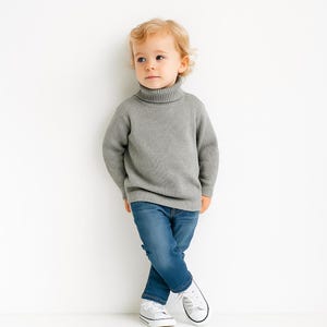 May include: A young child wearing a gray ribbed turtleneck sweater and blue jeans. The child is also wearing white sneakers with black trim. The child is standing in front of a white background.