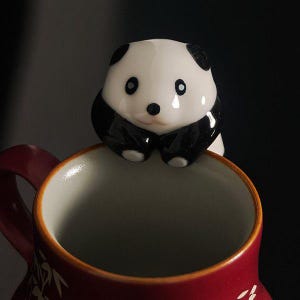 May include: A red ceramic mug with a white interior, topped with a panda bear figurine. The panda has black and white markings. The mug's handle is red, and the exterior has a decorative pattern.