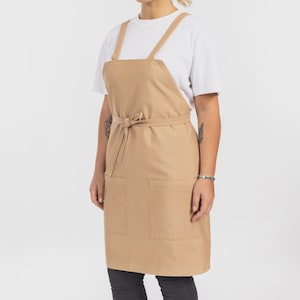May include: A tan apron featuring a cross-back strap design, a waist tie, and two large front pockets. The apron is worn over a plain white short-sleeved t-shirt and dark pants, providing a neutral and functional appearance for various tasks.