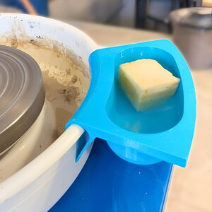 Könnte beinhalten: Eine Töpferscheibe mit einer blauen Zubehörschale, die einen gelben Schwamm enthält. Das Rad ist weiß und hat eine sich drehende Metalloberfläche. Die Schale ist am Rad befestigt und liegt auf einer blauen Oberfläche.