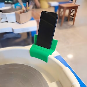 May include: A green plastic phone holder with the word "WACO" printed on it, holding a black smartphone. The holder is attached to the rim of a pottery wheel. The wheel is white with a blue stripe.