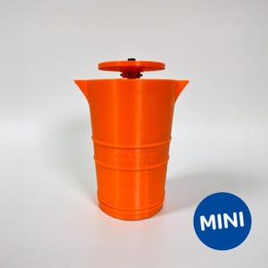 May include: Vibrant orange plastic cup with a circular top and a blue circle with the word "MINI" in white. The cup has a cylindrical shape with horizontal ridges and the word "PLAYGO" embossed on the side.