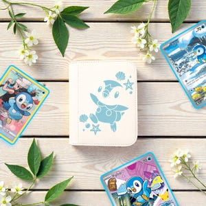 Pu&ograve; includere: Un raccoglitore per carte bianco con una grafica azzurra del personaggio Piplup circondato da piccole conchiglie e stelle, accompagnato da tre carte collezionabili che raffigurano lo stesso personaggio in varie scene colorate.