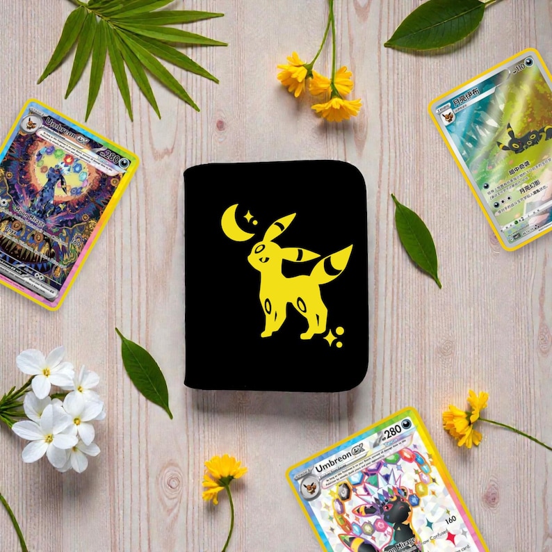 Pu&ograve; includere: Un portafoglio rettangolare nero con una grafica gialla del Pokemon Umbreon in piedi sotto una luna crescente e delle stelle. Il portafoglio &egrave; circondato da tre carte collezionabili e da diversi fiori gialli e foglie verdi su una superficie in legno.