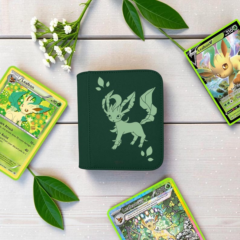 Pu&ograve; includere: Un raccoglitore per carte quadrato verde scuro con una silhouette verde chiaro del Pok&eacute;mon Leafeon sulla copertina, circondato da tre carte collezionabili Leafeon e un piccolo rametto di fiori bianchi con foglie verdi su una superficie in legno.