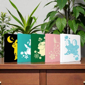 May include: A row of five colorful notebooks with animal-themed designs. The notebooks are black, teal, green, pink, and white. Each notebook features a different animal silhouette in a contrasting color, including a rabbit, frog, and duck.