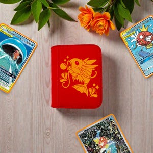 Pu&ograve; includere: Un raccoglitore per carte rettangolare rosso brillante con una grafica arancione stilizzata di un pesce con pinne e bolle. Il raccoglitore &egrave; circondato da tre carte collezionabili che raffigurano pesci e fiori arancioni con foglie verdi.