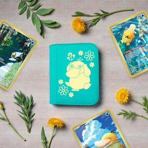 Pu&ograve; includere: Un raccoglitore per carte color verde acqua con una grafica giallo chiaro del personaggio Psyduck circondato da piccole icone di fiori e punti interrogativi, posizionato tra diversi fiori gialli e foglie verdi.