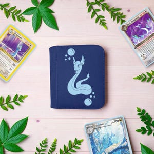 Pu&ograve; includere: Un raccoglitore per carte quadrato blu scuro con una silhouette azzurra della creatura serpentina Dragonair circondata da piccole bolle e stelle. Il raccoglitore &egrave; circondato da tre carte collezionabili che raffigurano la stessa creatura.
