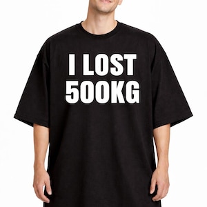 Può includere: T-shirt nero oversize con la scritta "I LOST 500KG" in lettere bianche in grassetto. La maglietta ha maniche corte e una vestibilità rilassata. Il modello indossa jeans blu.