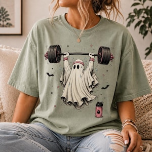 May include: A sage green short-sleeve t-shirt featuring a graphic of a white ghost wearing a pink headband and lifting a black barbell. The design includes small stars, bats, and a pink water bottle with a ghost icon printed on the front.