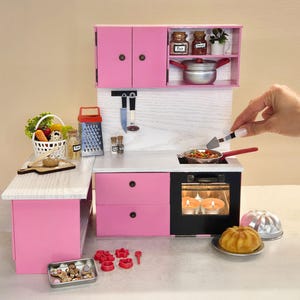 K&ouml;nnte beinhalten: Eine Miniatur-K&uuml;chenzeile in Rosa mit Herd, Schr&auml;nken und Zubeh&ouml;r. Das Set enth&auml;lt einen Korb mit Gem&uuml;se, eine Reibe und eine Pfanne mit Essen. Eine Hand verwendet einen Pfannenwender, um das Essen in der Pfanne zu wenden.