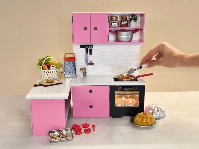 K&ouml;nnte beinhalten: Eine Miniatur-K&uuml;chenzeile in Rosa mit Herd, Schr&auml;nken und Zubeh&ouml;r. Das Set enth&auml;lt einen Korb mit Gem&uuml;se, eine Reibe und eine Pfanne mit Essen. Eine Hand verwendet einen Pfannenwender, um das Essen in der Pfanne zu wenden.