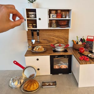 May include: Miniature kitchen set with white cabinets, a wooden countertop, and a black oven. Includes a red-handled pan, a cake, and various cooking utensils. The set is decorated with miniature food items and spices.