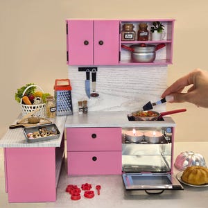 K&ouml;nnte beinhalten: Eine Miniatur-K&uuml;chenzeile in Rosa mit Herd, Schr&auml;nken und Zubeh&ouml;r. Das Set enth&auml;lt einen kleinen Topf, eine Reibe, einen Gem&uuml;sekorb und ein Backblech. Eine Hand h&auml;lt einen Spatel &uuml;ber einer Pfanne auf dem Herd.