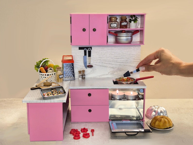 K&ouml;nnte beinhalten: Eine Miniatur-K&uuml;chenzeile in Rosa mit Herd, Schr&auml;nken und Zubeh&ouml;r. Das Set enth&auml;lt einen kleinen Topf, eine Reibe, einen Gem&uuml;sekorb und ein Backblech. Eine Hand h&auml;lt einen Spatel &uuml;ber einer Pfanne auf dem Herd.
