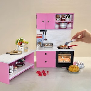 K&ouml;nnte beinhalten: Eine Miniatur-K&uuml;chenzeile in Rosa mit einer wei&szlig;en Arbeitsplatte und einem schwarzen Ofen. Das Set umfasst einen Herd, Schr&auml;nke und eine kleine Insel. Ein Korb mit Gem&uuml;se und ein Kuchen sind ebenfalls vorhanden.