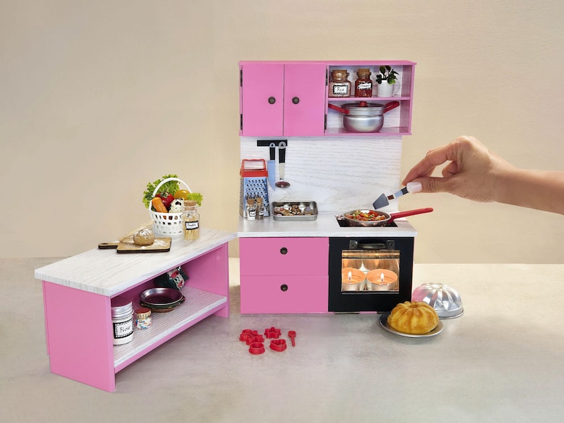 K&ouml;nnte beinhalten: Eine Miniatur-K&uuml;chenzeile in Rosa mit einer wei&szlig;en Arbeitsplatte und einem schwarzen Ofen. Das Set umfasst einen Herd, Schr&auml;nke und eine kleine Insel. Ein Korb mit Gem&uuml;se und ein Kuchen sind ebenfalls vorhanden.