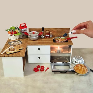 Puede incluir: Escena de cocina en miniatura con encimera y armarios de madera y blancos. Incluye una cesta de verduras, un rallador, ollas, sartenes y un pequeño horno con una vela encendida. Se muestra una mano agregando algo a una sartén en la estufa.