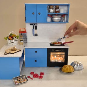 Könnte beinhalten: Eine Miniatur-Küche in Blau und Weiß mit Herd, Schränken und einer Arbeitsplatte. Das Set enthält eine Bratpfanne mit Essen, einen Korb mit Gemüse und Backutensilien. Eine Hand hält einen Spatel über der Pfanne.