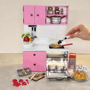 K&ouml;nnte beinhalten: Miniatur-K&uuml;chenset in Rosa mit Schr&auml;nken, Schubladen und Ger&auml;ten. Enth&auml;lt einen Herd, einen Backofen und Zubeh&ouml;r wie einen Gem&uuml;sekorb, Kochutensilien und einen Kuchen. Eine Hand wird gezeigt, die einen Spatel zum Kochen von Speisen in einer Pfanne verwendet.