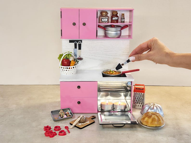K&ouml;nnte beinhalten: Miniatur-K&uuml;chenset in Rosa mit Schr&auml;nken, Schubladen und Ger&auml;ten. Enth&auml;lt einen Herd, einen Backofen und Zubeh&ouml;r wie einen Gem&uuml;sekorb, Kochutensilien und einen Kuchen. Eine Hand wird gezeigt, die einen Spatel zum Kochen von Speisen in einer Pfanne verwendet.