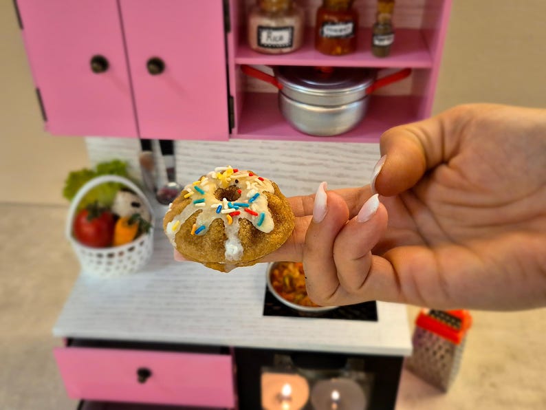 K&ouml;nnte beinhalten: Ein Miniatur-Gugelhupf mit wei&szlig;er Glasur und bunten Streuseln wird vor einer rosa-wei&szlig;en Puppenk&uuml;che gehalten. Die K&uuml;che verf&uuml;gt &uuml;ber einen Herd, Schr&auml;nke und einen kleinen Topf. Ein Korb mit Gem&uuml;se ist ebenfalls zu sehen.