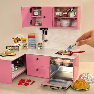 K&ouml;nnte beinhalten: Eine Miniatur-K&uuml;chenzeile in Rosa mit offenen Schr&auml;nken, einem Herd und einem Backofen. Das Set enth&auml;lt einen Korb mit Gem&uuml;se, Gew&uuml;rzgl&auml;ser und einen kleinen Topf. Eine Hand r&uuml;hrt mit einem Spatel Essen in einer Pfanne auf dem Herd um.
